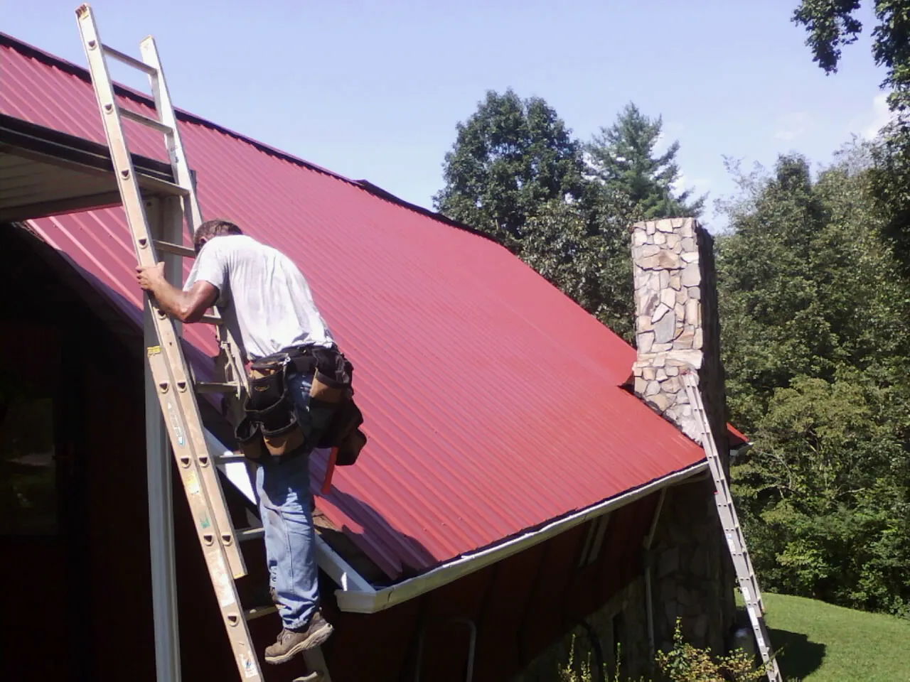 Roofer on a ladder beside a red metal roof and stone chimney in West Jordan